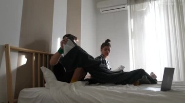 Yatak odalarında rahat bir anın tadını çıkaran genç bir çift rahat bir ortamda dizüstü bilgisayarlarına odaklanmış..