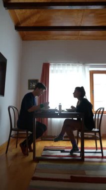 İki insanın keyifle yemek yiyip sohbet ettiği sıcak bir kahvaltı sahnesi.