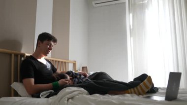 Genç bir adam yatağında rahat bir şekilde dizüstü bilgisayar kullanarak rahat ve rahat bir ev ortamı yaratıyor.