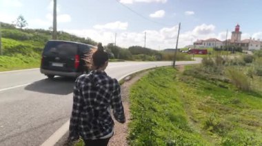 Modaya uygun genç bir kadın güneşli bir gökyüzünün altında büyüleyici bir deniz fenerinin önünden geçen büyüleyici bir yolda yürüyor.