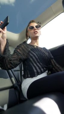 Siyah bluzlu ve güneş gözlüklü şık genç bir kadın zarif, lüks bir arabanın içinde selfie çekiyor.