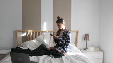 Genç bir kadın rahat bir şekilde yatağında oturuyor, akıllı telefonunu modern dekorlu, şık bir yatak odasında kullanıyor.