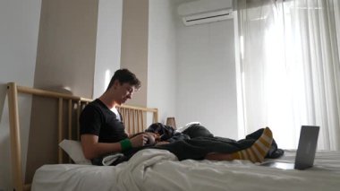 Genç bir adam rahat bir şekilde yatağında dizüstü bilgisayarını ve telefonunu parlak, güneşli bir yatak odasında kullanıyor.