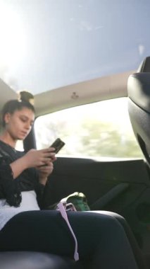 Genç bir kadın, hareket halindeki bir arabanın arka koltuğunda rahatça otururken akıllı telefonuna odaklanır.