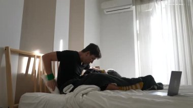 Genç bir adam rahat bir şekilde yatağında oturuyor, bilgisayarına odaklanmış, huzurlu bir rahatlama anının tadını çıkarıyor.