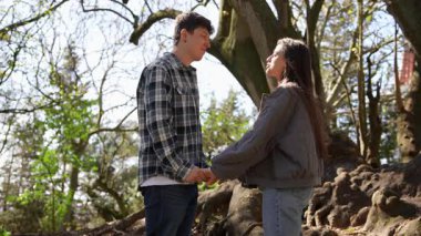 Genç bir çift doğada güzel bir anın tadını çıkarır, neşeyle gülümser ve yapraklı ağaçların altında el ele tutuşur.