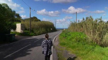 Genç bir kadın, yemyeşil ve parlak mavi gökyüzü ile çevrili güzel ve güzel bir yolda yürüyor.