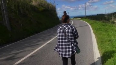 Bir kadın yemyeşil yemyeşil bir yolda geziniyor ve sakin bir manzarada araçların yanından geçiyor..