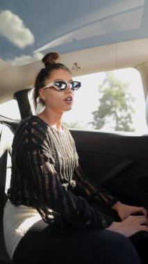 Şık bir genç kadın arabanın arka koltuğunda şık güneş gözlükleriyle birlikte şık kıyafetlerini sergiliyor.