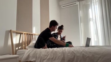 Bir çift, sıcak ve samimi bir anı paylaşırken dizüstü bilgisayarlarıyla yakın bir şekilde bir yatakta oturuyor.