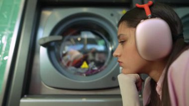 Renkli kıyafetlerinin çamaşır makinesinde dönüşünü seyreden bir kız. Güvenlik için kulak koruyucusu takıyor.