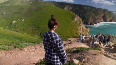 Genç bir kadın yüksek bir uçurumda duruyor, güneşli bir günde nefes kesen güzel kıyı manzarasına bakıyor.