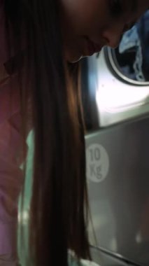 Bir kadın çamaşır makinesine çamaşır yüklemekle meşgul. Günlük hayattan basit ama anlamlı bir anı yakalıyor.