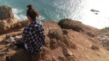 Dalgın bir kadın kayalık bir uçurumda huzur içinde oturuyor ve aşağıdaki büyüleyici okyanus manzarasına dikkatlice bakıyor.