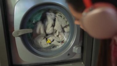 Genç bir insan modern bir makinede yıkanan kıyafetleri dikkatle izler, günlük çamaşırların rutinini gösterir.