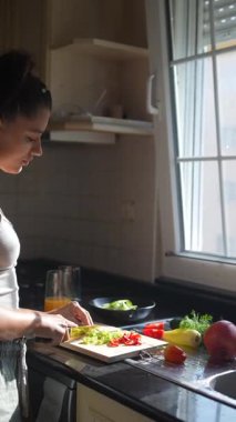 Bir kadın aydınlık, güneşli bir mutfakta taze sebzeler hazırlarken ferahlatıcı bir içkinin tadını çıkarıyor.