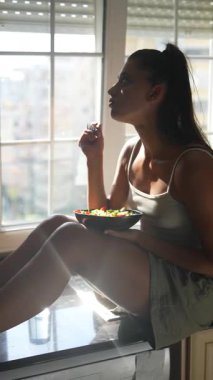 Pencerenin önünde oturan, taze ve renkli bir salatanın tadını çıkaran sağlıklı bir yaşam tarzını vurgulayan bir kadının silüeti.