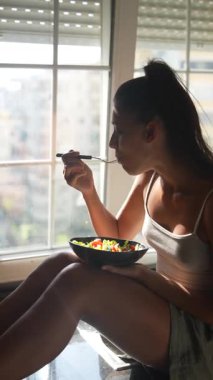 Genç bir kadının güneşli bir pencerede renkli ve canlı bir salatanın tadını çıkardığı huzurlu ve huzurlu bir sahne.