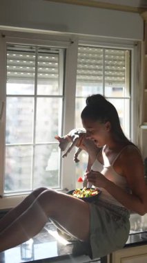 Bir kadın mutfak tezgahına oturmuş, güneş ışığıyla yıkanırken lezzetli ve sağlıklı bir yemeğin tadını çıkarıyor.