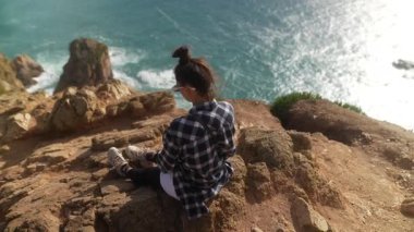 Huzurlu ve güzel bir an kayalık bir uçurumda sessizce oturan ve altındaki engin okyanusa bakan genç bir kız.