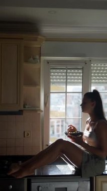 Genç bir kadın mutfak tezgahında rahat bir şekilde otururken doğal ışıkta canlı ve renkli bir salatanın tadını çıkarıyor.