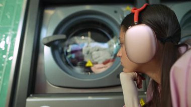 Bir kız çamaşır makinesinde dönen rengarenk çamaşırlarını izlemekten, kulaklık takmaktan ve anın tadını çıkarmaktan hoşlanır.