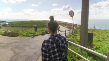 Bir insan canlı yeşillikler ve çarpıcı okyanus manzaralarıyla çevrili manzaralı bir sahil yolunda yürüyor.