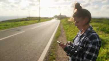 Genç bir kadın sessiz bir yolda duruyor, güneş batarken telefonunu kontrol ediyor, sakin bir atmosfer yaratıyor.