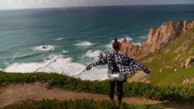 Bir insan uçurumun kenarında durmuş güneşli bir günde hafif esintinin ve nefes kesici okyanus manzarasının tadını çıkarıyor.