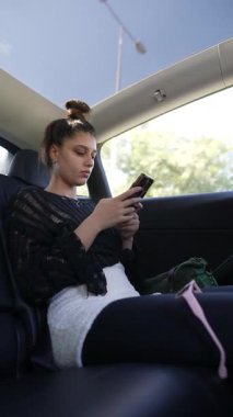 Genç bir kadın arabada oturuyor, akıllı telefonuna odaklanmış, modern yaşam tarzı ve teknoloji kullanımı sergiliyor..