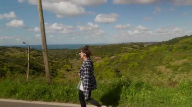 Genç bir kadın, bereketli tepeler ve okyanus manzaralarıyla çevrili, doğa güzelliğini kucaklayan manzaralı bir yolda yürüyor.