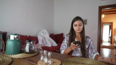 Akıllı telefonuna kendini kaptırmış genç bir kadın rahat bir yaşam alanında oturur ve modern bir yaşam tarzını yansıtır.