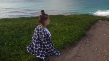 Genç bir kadın yemyeşil ve çarpıcı okyanus manzaralarıyla süslenmiş güzel bir sahil yolunda rahatça yürüyor.