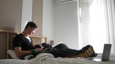 Genç bir adam dijital cihazlarla yatakta rahatlıyor, modern rahatlık ve rahatlık dolu rahat bir yaşam tarzı sergiliyor