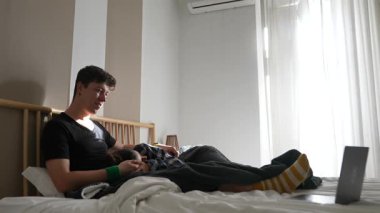 Genç bir adam yatağında rahat bir şekilde dizüstü bilgisayar kullanarak rahat ve rahat bir ev ortamı yaratıyor.