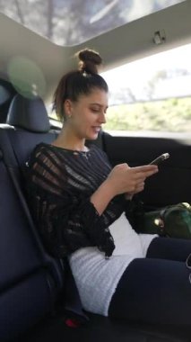 Genç bir kadın, bir arabanın arka koltuğunda rahatça otururken akıllı telefonuyla eğleniyor.