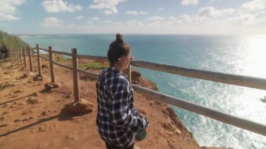 Genç bir kadın uçurumun kenarında duruyor, büyüleyici okyanus manzarasının tadını çıkarıyor, parlak ve güzel bir gökyüzünün altında.