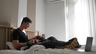 Genç bir adam rahat bir şekilde yatağında oturuyor, rahat bir ortam içinde mobil cihazıyla meşgul.