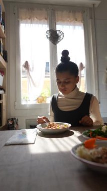 Genç bir kız, güneşli bir mutfak masasında sıcak bir atmosferde, canlı tabaklarla çevrili bir şekilde yemeğin tadını çıkarıyor.