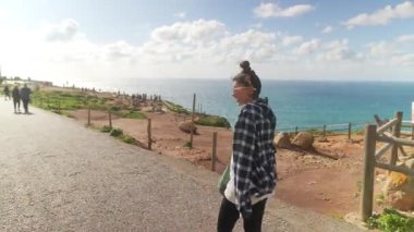 Genç bir kadın, bir arkadaşıyla güzel bir sahil yolunda neşeyle yürüyor, nefes kesici okyanus manzarasının tadını çıkarıyor.