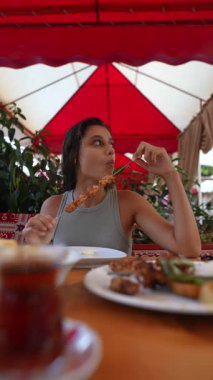 Bir kadın, parlak, renkli bir gölgeliğin altında büyüleyici bir restoranda şiş şiş eğleniyor.