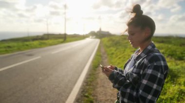 Genç bir kadın sessizce manzaralı bir yolun kenarında duruyor, telefonuna odaklanmış, nefes kesici manzarayla çevrili.