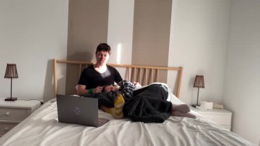 Genç bir insan, parlak bir yatak odasında yumuşak ve rahat battaniyelerle çevriliyken dizüstü bilgisayarıyla mutlu mesut meşgul olur.