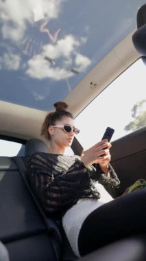 Şık ve şık genç bir kadın modern bir arabanın içinde rahatlarken akıllı telefonuyla mutlu mesut meşgul oluyor.