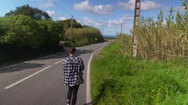 Bir insan, berrak mavi gökyüzünün altında yemyeşil bir güzellikle çevrili huzurlu bir yolda rahatça yürür.