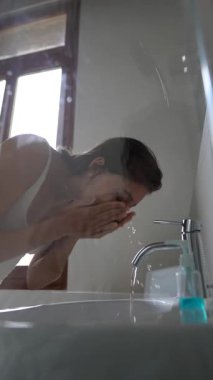 Genç bir kadın, aydınlık ve modern bir banyo ortamında dururken ferahlatıcı bir yüz yıkamanın tadını çıkarıyor.