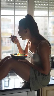 Bir kadın mutfak tezgahında oturmuş renkli ve sağlıklı bir salatanın tadını çıkarıyor. Parlak, davetkar mutfağında.