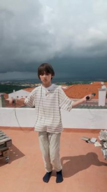 Genç bir çocuk neşeyle çatıda ip atlıyor, dramatik bir bulutla çevrili, gençlik ruhunu sergiliyor.