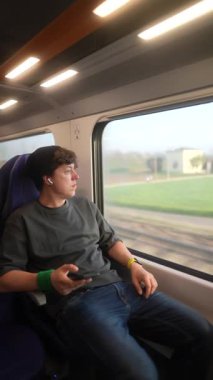 Genç bir insan düşünceli bir şekilde hareket halindeki bir trenin penceresinden dışarı bakar.