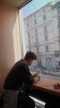 Genç bir insan akıllı telefonuna dalmış, penceresinin dışındaki kentsel manzarayı inceliyor.
